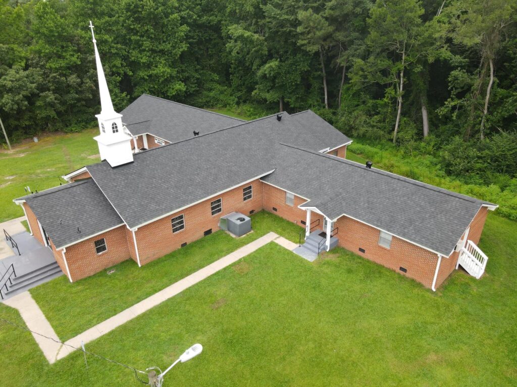 Aerial view of a red-brick church with a white steeple and black roofing surrounded by lush green trees. The building features multiple connected sections with white framed windows and concrete pathways leading to staircases at the entrances.