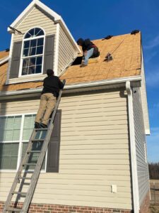 Two people are working on the roof of a house. One person is on a ladder, while the other is kneeling on the roof, performing precise roofing tasks on the wooden sheathing. The sky is clear and blue, and the house has beige siding and large windows.