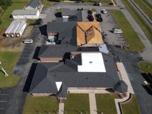 An aerial view of a large building with multiple sections and different roof types, including new shingles installed by Halo Roofing. One section has a tan underlayment, another features a white material. Surrounding areas include parked vehicles, green grass, and nearby roads.