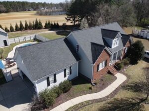 Aerial view of a suburban home with a gray roof by Halo Roofing and partially brick facade. The property features a neatly landscaped yard, a curved sidewalk, an attached garage, and an in-ground pool in a fenced backyard. Surrounded by trees and a field in the background.