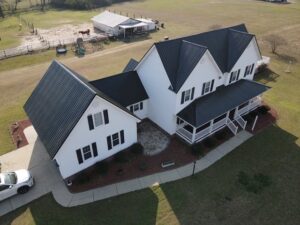 Aerial view of a large, white, two-story farmhouse with sleek black roofs by Halo Roofing and a wraparound porch. There is a circular driveway with a parked car, several outbuildings, and a fenced area with a horse and farm equipment in the background, surrounded by green fields.