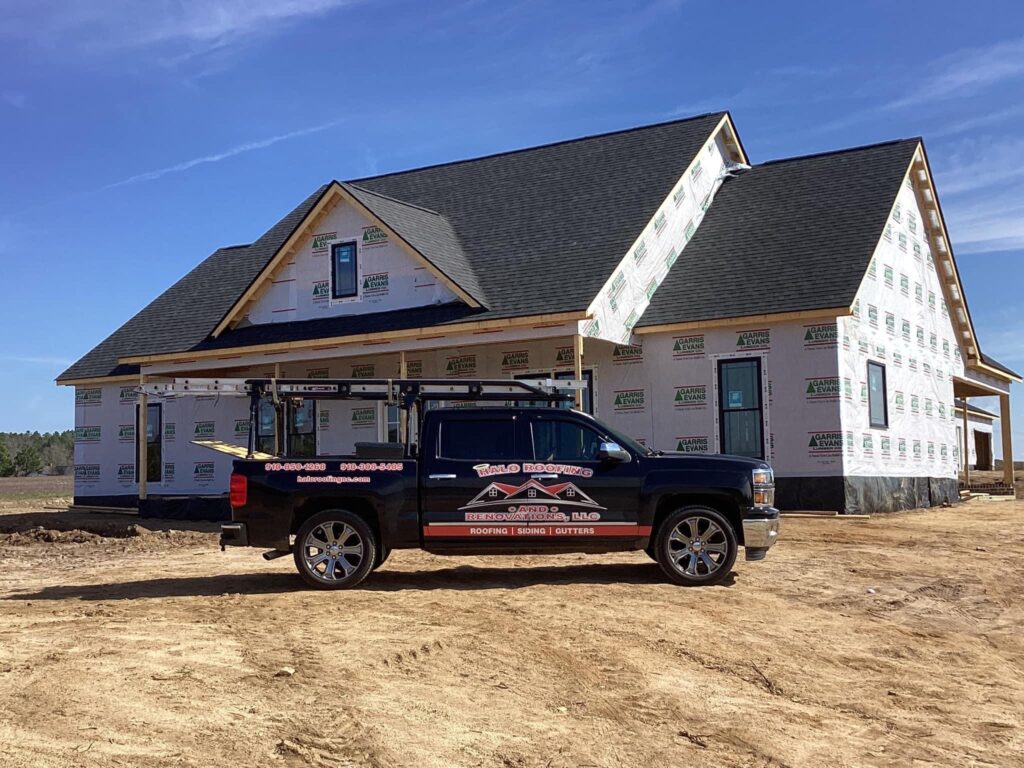 A newly constructed house with a white exterior wrap and a sloped roof is partially finished. A black pickup truck with advertising for Halo Roofing and Renovations LLC is parked in front on a dirt driveway. The sky is clear and blue.