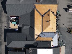 An aerial view of a building undergoing roof construction by Halo Roofing. Part of the roof is covered in shingles, while the other part is exposed wood sheathing. Several workers are actively roofing. Surrounding areas include cars and construction materials.