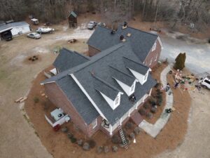 Aerial view of a brick house with white trim and a dark gray gabled roof being worked on by several roofing experts. The house has a large front porch, and the surrounding area features landscaping with bushes and mulch, a driveway, and some additional buildings.