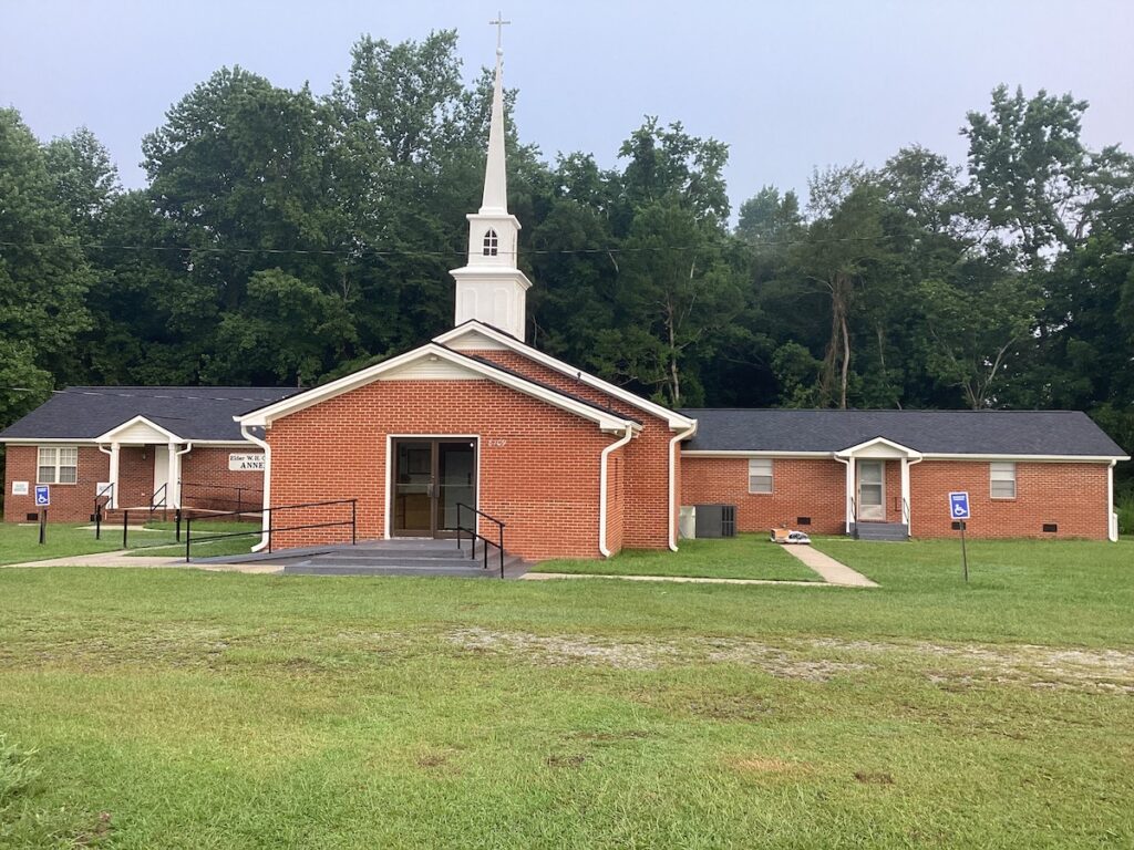 A small red-brick church with a white steeple and cross, featuring roofing, flanked by two similarly styled buildings. The church has a ramp for accessibility. There are trees and greenery in the background and a grassy area in the foreground.