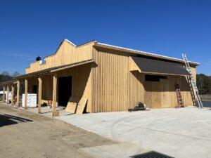 A partially constructed wooden building stands on a concrete foundation under a clear blue sky. Wooden panels, ladders, and various construction materials are scattered around the site as workers prepare for the installation of roofing.