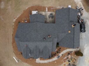 An aerial view captures a large, dark-roofed house surrounded by brown landscaping. Workers from Halo Roofing are visible on the roof, with construction materials and equipment scattered around the property, including a truck and various items on the ground.