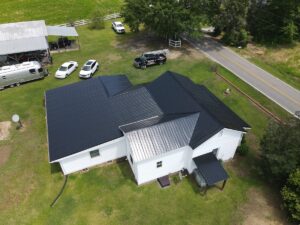 Aerial view of a white house with roofing on a grassy lot. The property includes multiple cars parked, a driveway, and a carport with a trailer. There is a road in the background with trees and a fence visible.