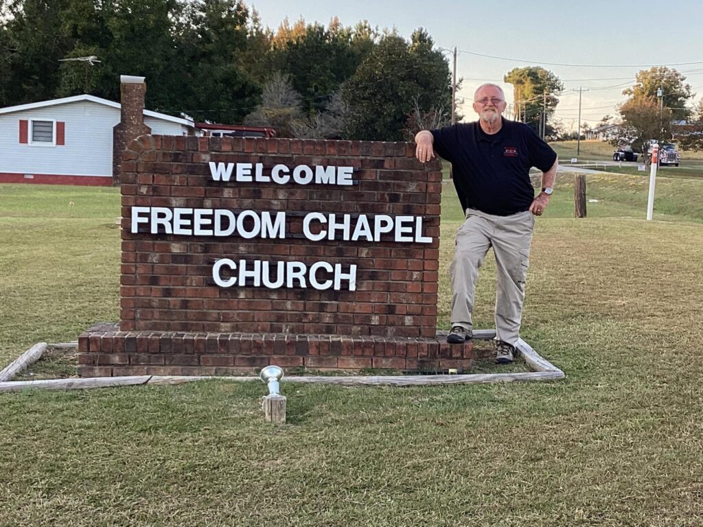 A person is leaning against a brick sign that reads "WELCOME FREEDOM CHAPEL CHURCH." They are outside on a grassy area with a small white building in the background, featuring roofing. Trees and a road are also visible in the distance.