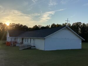 A white, single-story church building with a cross on the roofing stands in a grassy field. The sun is setting behind the trees in the background, casting a warm glow on the scene. A wheelchair ramp is visible on the side of the church.