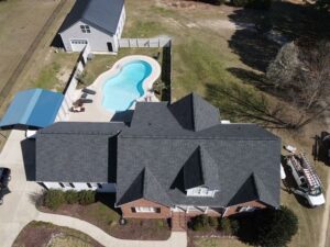 Aerial view of a house with a dark gray roof by Halo Roofing and red brick exterior. The property includes a well-maintained backyard with an irregularly shaped pool surrounded by a white fence. Adjacent to the pool is a deck with lounge chairs and a blue-roofed carport near a separate garage.