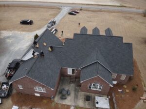 Aerial view of a large red-brick house with several workers from Halo Roofing on the roof, repairing shingles. The house has multiple gables and dormer windows. Construction vehicles and materials are scattered around the property, with a road visible in the background.