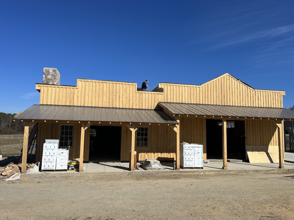 A wooden building under construction stands against a clear blue sky. The building features a porch, two large openings, and several crates and construction materials scattered around. A person is visible working on the roofing near the chimney.