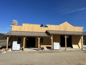 A wooden building under construction stands against a clear blue sky. The building features a porch, two large openings, and several crates and construction materials scattered around. A person is visible working on the roofing near the chimney.