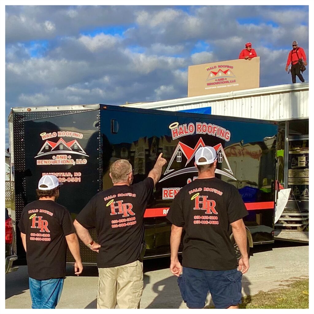 Three men in Halo Roofing branded shirts stand near a company truck, pointing at two workers positioning a large roofing sign on the roof of a building. The truck features the Halo Roofing logo, while one worker on the roof holds the sign in place.