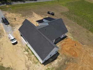 Aerial view of a one-story house with dark roofing and white exterior, set on a dirt and grass-covered plot. There is a white van and a black truck with ladders on the side. A solar panel setup is seen adjacent to the house on the roof.