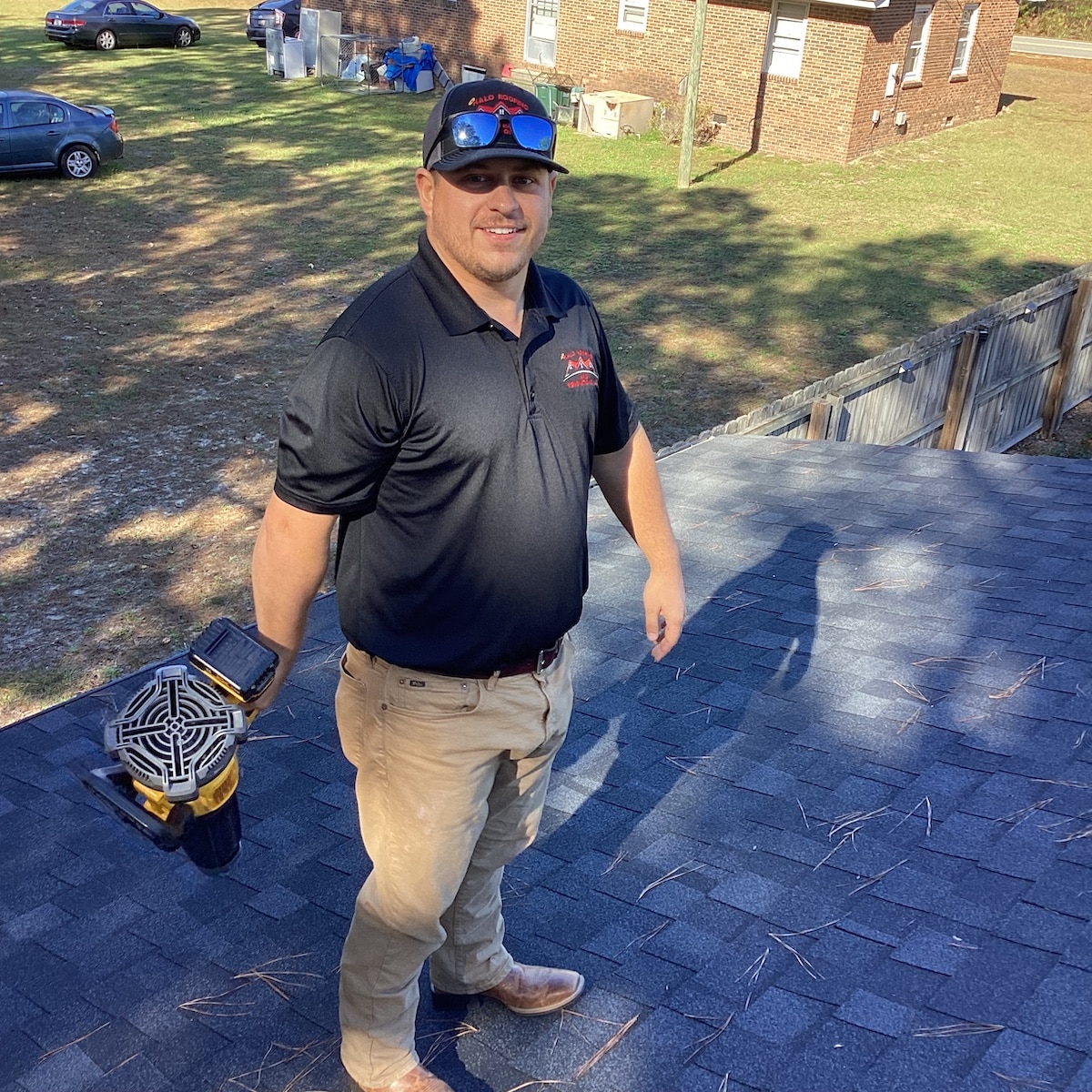 A man in a black polo shirt and khaki pants holds a power tool while standing on a shingled roof, showcasing his roofing skills. He smiles at the camera. In the background, a yard with a wooden fence, two vehicles, and brick buildings are visible. Pine needles are scattered on the roof.