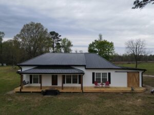 A single-story house with white walls and a black roofing featuring a covered wooden porch. The porch has a black swing on the left and two red chairs with a small table on the right. There are trees and an open field in the background under an overcast sky.