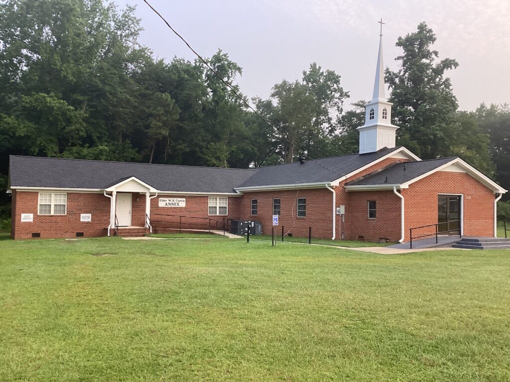 A small red-brick church with a white steeple and roofing sits in a grassy area surrounded by trees. A sign reading "ANNEX" is displayed near the entrance, and a ramp leads up to the door. The church has a simple rectangular design with a few small windows.