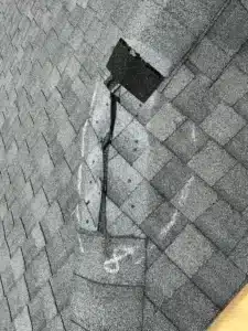 A close-up view of a gray shingled roof with visible damage. Several shingles are missing or displaced, exposing the underlying black material and nails. The area is marked with chalk circles, indicating the extent of the damage, suggestive of a much-needed Halo Roofing inspection.