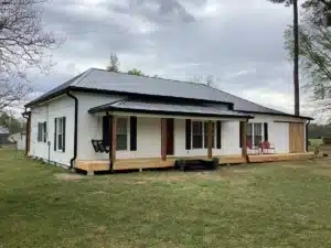 A single-story white house with a black Halo Roofing-covered roof and trim has a covered front porch with a wooden swing, two red chairs, and a side yard. The sky is overcast. The house features a mix of vertical and horizontal siding and multiple windows, including a set of French doors.