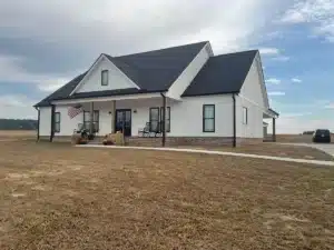 A white, single-story house with halo roofing and black trim, a spacious front porch, and a gable roof. An American flag is displayed on the porch. The house is situated in an open area with dry grass. A black car is parked on the driveway to the right. The sky is partly cloudy.