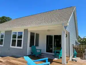 A light gray single-story house with a brown shingle roof by Halo Roofing features a backyard patio. The patio has two light blue Adirondack chairs, white walls, and a ceiling fan. The patio leads out to a wooden deck with additional seating. The sky is clear and blue.