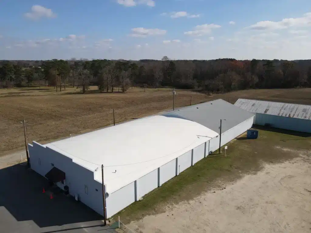 Aerial view of a long, white-roofed building, showcasing exquisite roofing, surrounded by open fields with some trees in the background. A smaller, darker-roofed structure is attached to the side of the main building. The sky is clear with some scattered clouds.