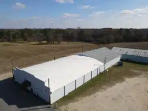 Aerial view of a long, white-roofed building, showcasing exquisite roofing, surrounded by open fields with some trees in the background. A smaller, darker-roofed structure is attached to the side of the main building. The sky is clear with some scattered clouds.