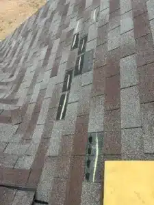 A roof with several missing shingles, leaving exposed patches that show underlying materials. Some shingle sections have been replaced by Halo Roofing, but the new shingles appear slightly different in texture and color. A flat, rectangular, yellow object is visible at the bottom right.