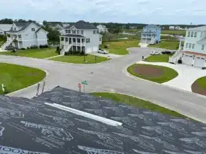 A view from a rooftop under construction with roofing underlayment installed reveals a residential neighborhood with several two-story homes, green lawns, and scattered trees. In the background, a body of water and more greenery stretch under a partly cloudy sky.