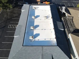 Aerial view of a large rectangular building with a flat, white roof and grey roofing around the perimeter. The roof has multiple ventilation units and other equipment installed. Surrounding areas include a parking lot with several parked vehicles and nearby structures.