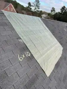 A large gray tarp is securely fastened over a section of a shingled roof on a residential home, as part of an ongoing roofing project. The tarp covers part of the roof, and “6 x 10” is written on the shingles. Trees and additional houses are visible in the background.