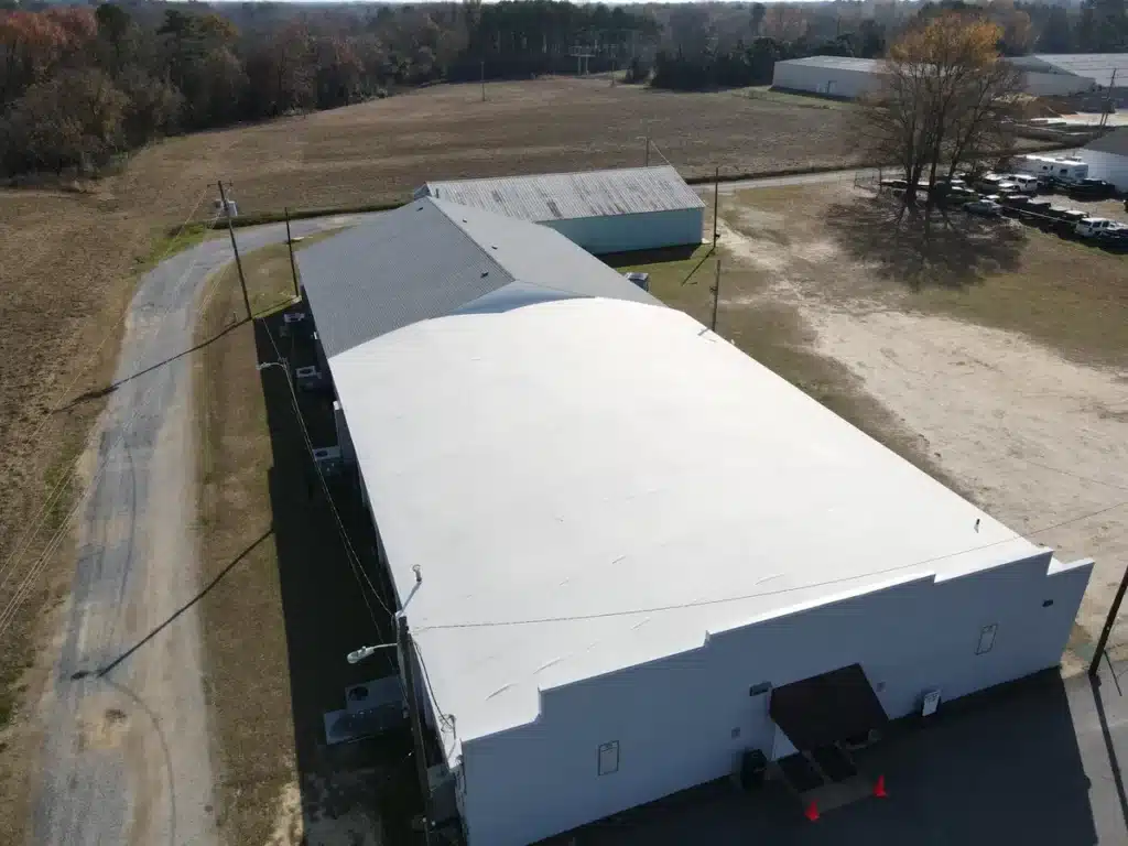 Aerial view of a large white warehouse building alongside a paved road. The surrounding area includes an open field and additional structures, with a few trees on the perimeter. Some vehicles are parked on one side of the warehouse.