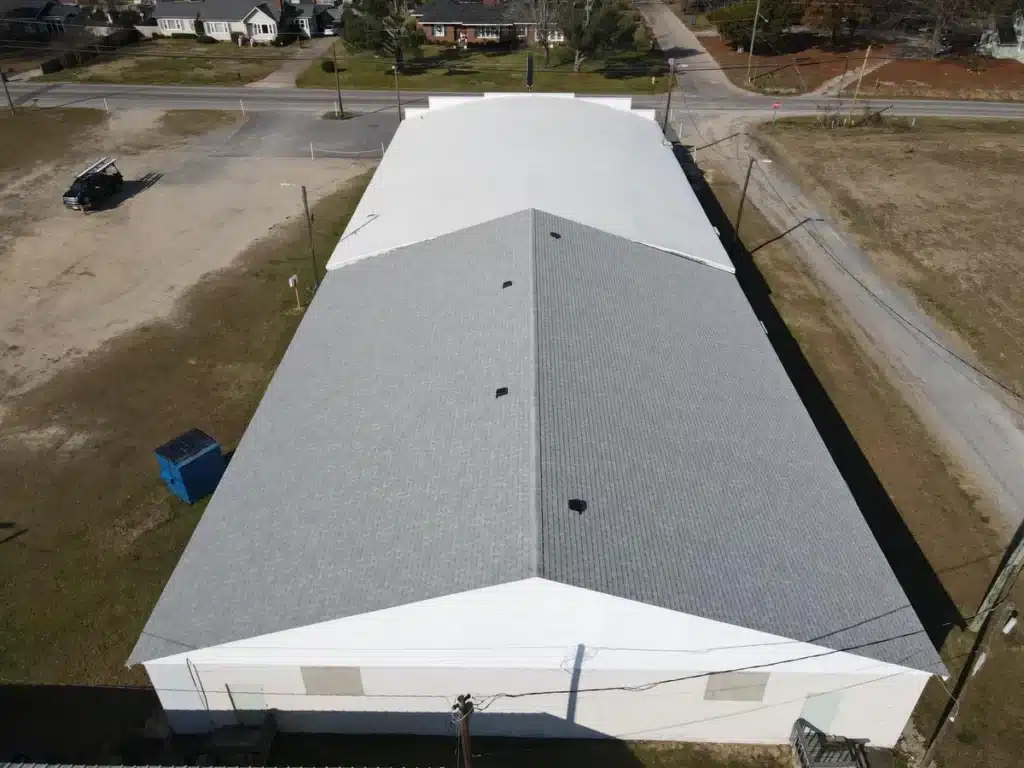 Aerial view of a large industrial building with a sleek, white roofing system, situated at the intersection of two roads with minimal traffic. The building is surrounded by grassy areas, a few smaller structures, and parked vehicles. The surrounding neighborhood features houses and trees.