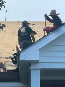 Two men work on a rooftop. One man is using a tool on the roof, while the other stands nearby. Both wear hats, and one has a t-shirt with a smiley face on the back. The roof is stripped to the wood, and roofing debris from Halo Roofing is scattered around.