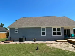 A single-story house with light blue siding and a sloped roof featuring halo roofing. The house has several windows and a backyard with a partially grassy lawn and patio area. Outdoor furniture is placed on the patio, and there is an air conditioning unit on the left side of the house.