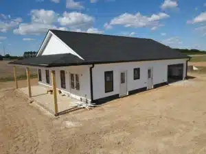 A single-story white house with black trim is under construction in a rural setting. It features a covered front porch supported by wooden posts and newly installed roofing. The property is surrounded by dirt and a partly cloudy blue sky overhead.