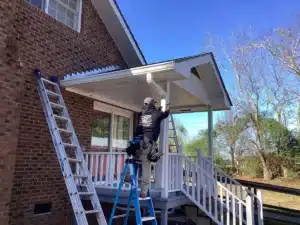 A person in work gear from Halo Roofing stands on a blue ladder, adjusting a gutter on the roof of a covered porch attached to a brick house. Nearby, another ladder leans against the house. The area is surrounded by trees under a clear blue sky.