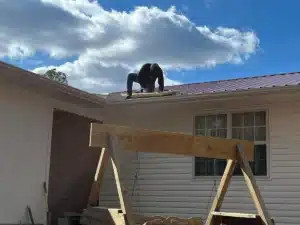 A person is kneeling on the sloped metal roof of a white house, holding a tool and working near the edge. Below them is a wooden porch swing, and the sky above is blue with scattered clouds. Their focused efforts on halo roofing ensure every detail is attended to.