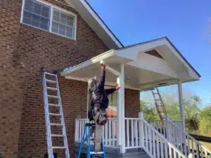 A worker wearing safety gear stands on a ladder, installing or repairing the halo roofing of a covered porch attached to a brick house. Two more ladders lean against the porch and house. The sky is clear and blue, with some greenery visible in the background.