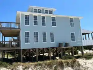 A light blue, two-story beach house with multiple tall windows is elevated on wooden stilts above sandy terrain with some greenery. A spacious wooden deck extends from the left side of the house, under a well-maintained roofing. The sky is clear and bright blue.