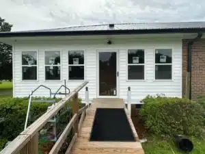 A white house with several windows, some featuring stickers, and a central glass door. A wooden ramp with handrails leads up to the door. Surrounding greenery includes hedges and potted plants. The dark-colored halo roofing beautifully contrasts with the light siding.