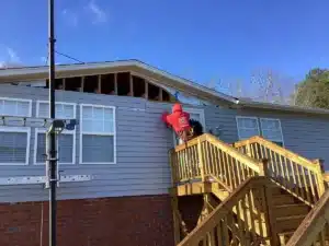 A worker in a red jacket stands on a wooden staircase, repairing the siding of a house with gray panels and exposed wooden beams near the roof. The house, featuring two white-framed windows and a brick foundation, is bathed in the clear blue sky—symbolic of Halo Roofing's commitment to excellence.