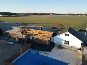 An aerial view of workers from Halo Roofing installing a new roof on a single-story house in a rural area. The roof is stripped down to the wood, and the surrounding area features fields, a swimming pool with a blue cover, and a small shed attached to the main house.