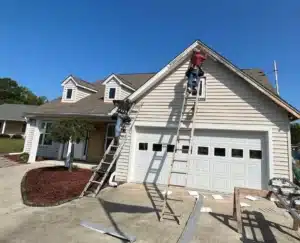 Two workers on ladders, from Halo Roofing, install siding on the exterior of a beige, two-story house with a double garage. The driveway is paved and there is a small garden with mulch and a tree to the left of the entrance. The sky is clear and blue.