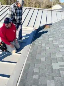 Two workers are on a roof, near the area where a metal roof section meets an asphalt shingle section. One is seated wearing a red hoodie and a cap, and the other is standing in a gray plaid shirt and cap. They appear to be inspecting or discussing the roofing work.