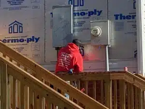 A worker in a red hoodie stands on a wooden deck, installing or inspecting electrical equipment on the exterior wall of a house under construction. The wall is covered with Thermo-ply sheathing, and an electrical meter box is visible above the halo roofing installation.