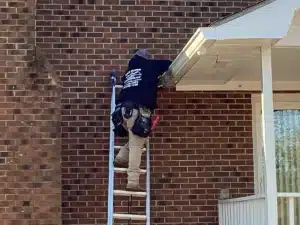 A person wearing a black shirt and khaki pants stands on a ladder, working on the fascia of a brick house. With tools in their belt, they’re positioned under the edge of the white roof near a downspout, diligently attending to roofing tasks.