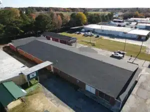 Aerial view of a small industrial or office complex featuring several connected one-story buildings with Halo Roofing, alongside a few standalone structures. The complex is surrounded by parking areas, trees, and a mix of grassy and paved areas. Nearby buildings and roads are visible in the background.